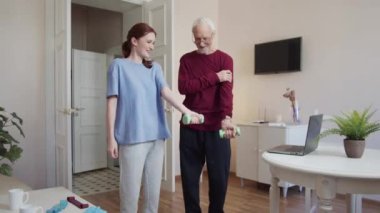 Genç bir kız, bir emeklinin dambıllarla egzersiz yapmasına yardım eder. Aynı anda yük kaldırıyorlar. Yüksek kalite 4k görüntü