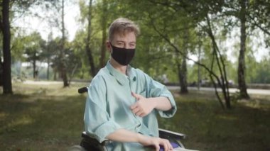 Kamu parkında, mavi gömlekli ve koruyucu maskeli engelli beyaz bir adam tekerlekli sandalyede oturmuş kameraya bakıyor. Yüksek kalite 4k görüntü