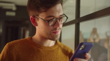 Yakışıklı genç bir öğrenci pencerenin yanında duruyor, elinde bir telefon tutuyor ve internette ilginç bir şey okuyor. Yüksek kaliteli FullHD görüntüler