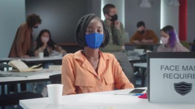 Genç bir Afrikalı profesör mavi koruyucu bir maske takmış sınıftaki öğrencileriyle kameraya bakıyor. Yüksek kalite 4k görüntü