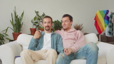 Lgbt genç yakışıklı adamlar kanepede birlikte oturuyorlar, el ele tutuşuyorlar ve kameraya bakıyorlar. Yüksek kalite 4k görüntü