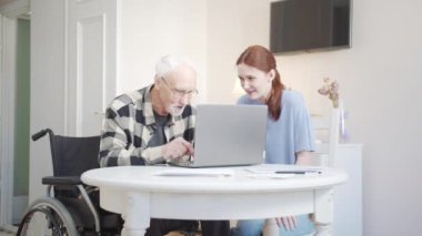 Sosyal servisten bir kız astına yardım ediyor. Tekerlekli sandalyedeki yaşlı bir adama laptopu nasıl kullanacağını söylüyor. Yüksek kalite 4k görüntü