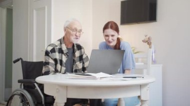 Bir adam dizüstü bilgisayarın önünde tekerlekli sandalyede oturuyor. Torunuyla birlikte, bir dizüstü bilgisayar kullanarak akrabalarıyla iletişim kuruyor.