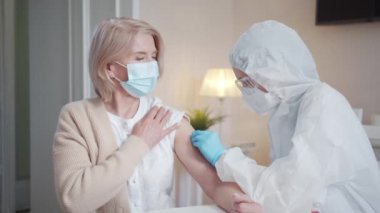 Aşıyı evde yapacağız. Tıbbi maskeli bir hemşire yaşlı bir kadına koronavirüs aşısı enjekte ediyor. Yüksek kalite 4k görüntü