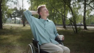 Tekerlekli sandalyedeki yetişkin bir beyaz adam parkta oturuyor ve havanın tadını çıkarıyor, içiyor ve konuşuyor. Yüksek kalite 4k görüntü