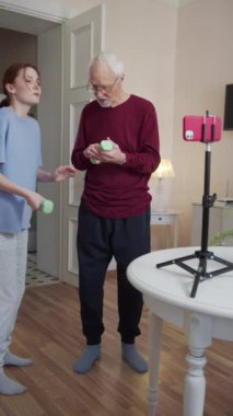 Kadın antrenör, dedeye dambılı nasıl kullanacağını söyler ve gösterir. Masanın önünde tripodun üzerinde bir akıllı telefon var.