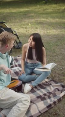 Sitting on the plaid in a public park, a young asian girl with long dark hair is reading a book for her disabled friend in a blue shirt. High quality FullHD footage