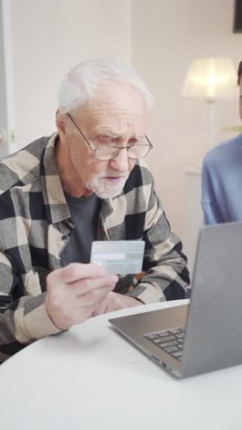 年金受給者は 店舗のウェブサイトに銀行カード番号を入力します 彼はラップトップを使っている 若い女性が隣に座って彼を助けます 高品質のフルHd映像 — ストック動画