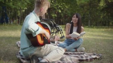 Elinde gitar olan engelli genç bir adam ekose üzerinde oturuyor ve kız arkadaşı kitap okuyup gülümserken şarkılar söylüyor. Yüksek kaliteli FullHD görüntüler