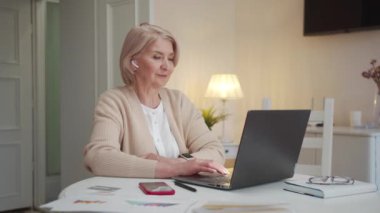 Bir kadın masadaki geniş bir odada oturuyor. O bir dizüstü bilgisayarda yazıyor. Kablosuz kulaklık kullanıyor. Akıllı saatiyle saati izliyor ve düğmesine basıyor. Yüksek kalite 4k görüntü