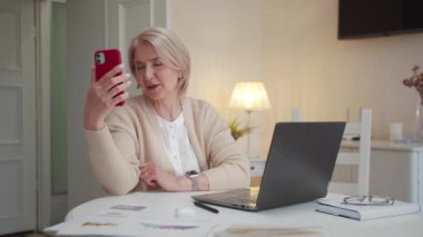  Yaşlı bir kadın büyük beyaz bir odada bir masada oturuyor. Akıllı telefon kullanarak görüntülü konuşma yapıyor. Müdahaleciyle vedalaştı ve telefonu kapattı. Yüksek kalite 4k görüntü