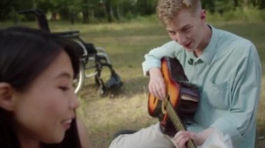 Halk parkında ekose üzerinde oturan Asyalı genç bir kız erkek arkadaşına gitar çalarak kitap okuyor ve ikisi de eğleniyor ve gülümsüyor. Yüksek kaliteli FullHD görüntüler