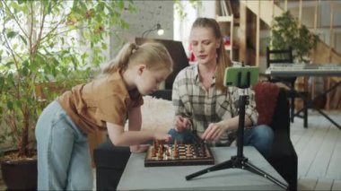 Anne ve kızı oturma odasında satranç oynuyorlar. Kız tahtaya beyaz parçalar koyuyor. Telefon kamerasını ayarlayan ve tripodu çeviren kadın. Yüksek kalite 4k görüntü