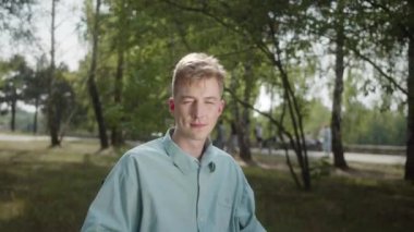 Tekerlekli sandalyedeki genç beyaz engelli bir adam parkta oturuyor ve ciddi ciddi kameraya bakıyor. Yüksek kalite 4k görüntü