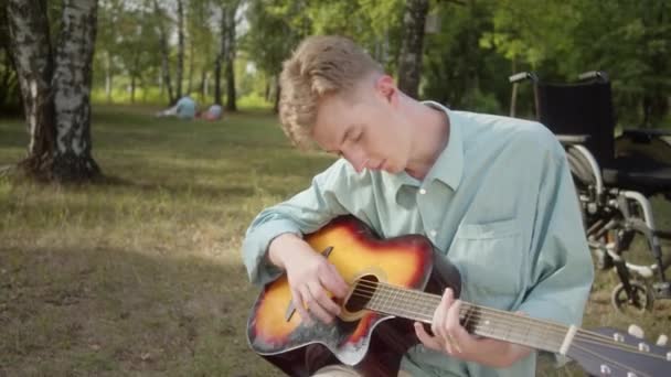 Młody Niepełnosprawny Blondyn Siedzi Kratę Parku Gra Zawodowo Gitarze Wysokiej — Wideo stockowe