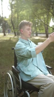 Engelli, sarışın bir adam elinde fincanla tekerlekli sandalyesinde oturuyor ve parkta fotoğraf çekiyor. Yüksek kaliteli FullHD görüntüler
