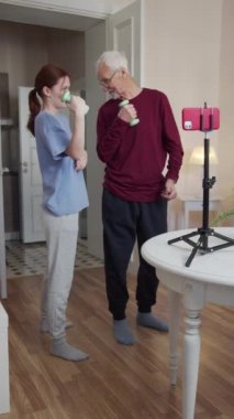 Yaşlı bir adam ve kızı birlikte spor yapıyorlar. Telefondaki talimatlara bakıyorlar ve halterleri senkronize olarak kaldırıyorlar. El değiştiriyorlar.