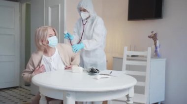 Koruyucu kıyafetli bir doktor hastadan ceketini çıkarmasını ve oturmasını istiyor. Hademe yaşlı bir kadının sırtına steteskop dayadı ve yüksek kaliteli 4K görüntüler dinledi.
