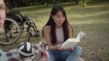 Kamu parkında ekose üzerinde oturan genç engelli bir adam, kız arkadaşı kitap okuyup gülümserken gitar çalıyor. Yüksek kaliteli FullHD görüntüler