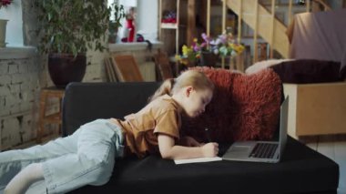 Küçük bir kız çalışma defterine yazıyor. Kanepede yüzüstü yatar ve dizüstü bilgisayara bakar. Yüksek kalite 4k görüntü