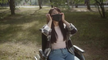 Uzun saçlı tatlı, engelli bir Asyalı kız koruyucu bir yüz maskesi çıkarıyor ve gülümsüyor. Yüksek kaliteli FullHD görüntüler