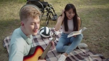 Sitting on the plaid in the public park, a young disabled man is playing guitar while his girlfriend is reading a book and enjoying the performance of her boyfriend . High quality FullHD footage