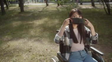 Çekici, neşeli Asyalı bir bayan halka açık bir parkta tekerlekli sandalyede oturuyor ve virüs salgını, salgın hastalık ve bulaşıcı hastalıkların önlenmesi için sağlık maskesi takıyor.