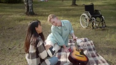 Kamu parkında ekose üzerinde oturan engelli bir genç kız arkadaşına gitar çalıp şarkı söylemeyi teklif ediyor. Yüksek kaliteli FullHD görüntüler