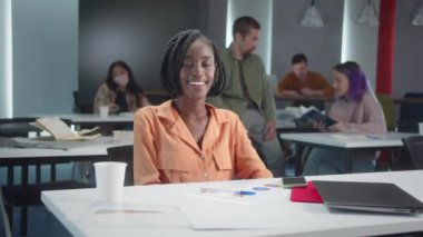 Sınıftaki masada oturan Afrikalı genç bir öğrenci kameraya bakıyor ve gülümsüyor, yavaş çekimde yakın çekim. Yüksek kaliteli FullHD görüntüler