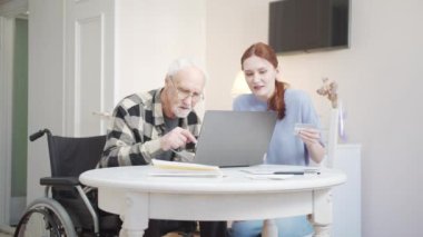Torunu dedesine yardım ediyor. Tekerlekli sandalyede oturan bir adam laptopla satın almaya çalışıyor. Yüksek kalite 4k görüntü