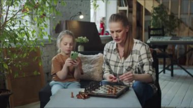 Bir kadın satranç taşlarını tahtaya yerleştirir. Kızı kanepede yanında oturuyor, canı sıkkın ve akıllı telefonunu kullanıyor. Yüksek kalite 4k görüntü