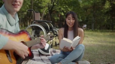 Sitting on the plaid in the public park, a young disabled man is playing guitar while his girlfriend is reading a book and enjoying the performance of her boyfriend . High quality FullHD footage