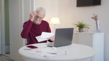 The pensioner is engaged in paper work. He sends bills and tax receipts through a laptop. He looks confused. High quality 4k footage