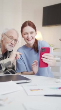 The woman and her a gray haired father in glasses uses the phone to communicate with relatives via video communication. They greet the interlocutor and wave their hand
