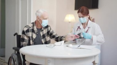 A female doctor sits at a table with an elderly man in a wheelchair. She puts a tonometer on his arm and begins to measure the pressure. High quality 4k footage