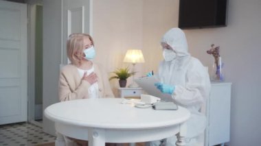 A doctor in a protective suit fills out a questionnaire about the patients physical condition. Woman complaining about heart problems. High quality 4k footage