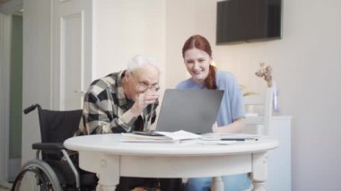A woman and a man are sitting at a table in a spacious apartment. Woman showing disabled person in wheelchair how to use laptop for communication