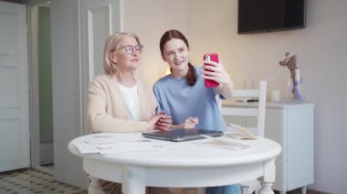 A woman and an old lady are sitting at a table in a large room. They are talking via video call. They greet the interlocutor and communicate nicely. High quality 4k footage