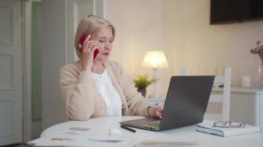 The pensioner is sitting at her desk. She is surrounded by papers and documents. An old woman communicates with a colleague on the phone and works at a computer, High quality 4k footage