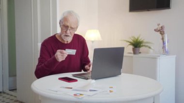 Grandfather uses a laptop to pay for services on the Internet. He carefully examines the credit card and drives the card number into the laptop.. High quality 4k footage