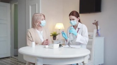 A woman in a white coat and with a stethoscope around her neck tells a pensioner about vaccination. The doctor offers two vaccines and asks to make a choice. High quality 4k footage