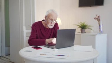An elderly man is having difficulty using a laptop. He is typing on the keyboard and looks flustered, High quality 4k footage