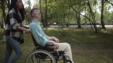 Genç ve güzel Asyalı bir kız parktaki tekerlekli sandalyede erkek arkadaşını yuvarlıyor ve Trey birbirleriyle konuşuyorlar. Yüksek kalite 4k görüntü