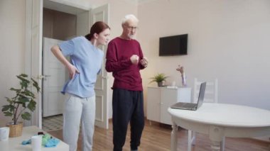 Kadın bir antrenör, yaşlı bir adama vücudunu eğip kollarını sallamasında yardım eder. Bilgisayar kullanarak video dersleri izliyorlar. Yüksek kalite 4k görüntü