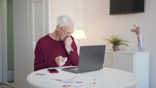 老人はノートパソコンの前のテーブルに座っている 彼はケースからイヤホンを取り出し 彼の耳に入れます 彼はコンピュータ上で曲をオンにし 陽気に一緒に歌い始めます — ストック動画