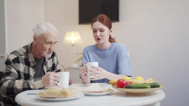 若い女性と車椅子の高齢者がテーブルに座り お茶を飲みます 果物やペストリーがテーブルの上にあります 人々はよく話す 高品質4K映像 — ストック動画