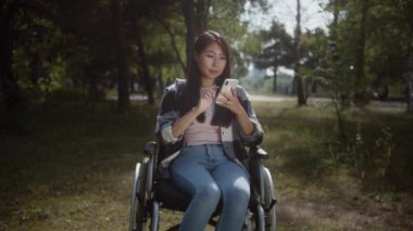 Güzel bir kız halka açık bir parkta tekerlekli sandalyede oturuyor ve cep telefonunun ekranına bakıp interneti arıyor ve gülümsüyor. Yüksek kalite 4k görüntü