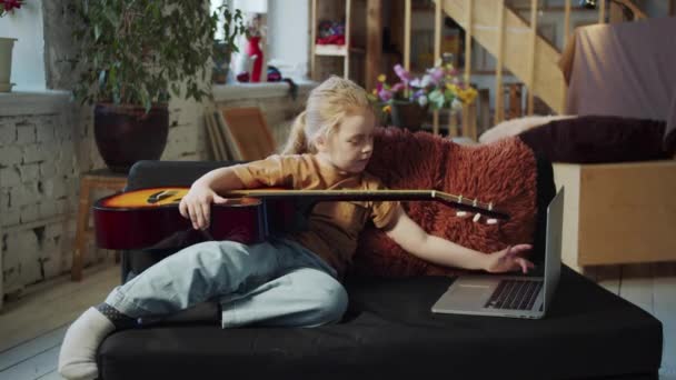 孩子坐在沙发上 手里拿着一把吉他 她上网查找有关这个工具的信息和一个教程 高质量的4K镜头 — 图库视频影像