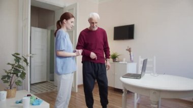 Yaşlı bir adam ve bir öğretmen birlikte fizik tedavi egzersizleri yapıyorlar. Bir kadın dizüstü bilgisayarda video dersi açar. Kol egzersizi yapıyorlar. Yüksek kalite 4k görüntü