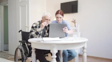 Bir kadın, tekerlekli sandalyedeki bir adama modern tisenolojiyi nasıl kullanacağını açıklar. Kablosuz kulaklıklarını laptoplarına taktılar ve müzik dinlediler. Büyükbaba mutlu ve şaşırdı. Yüksek kalite 4k